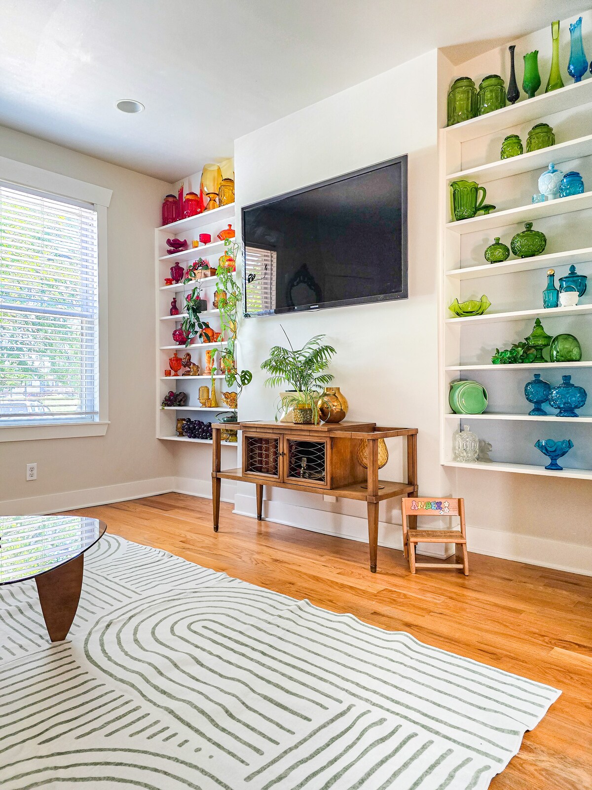 A bright living area showcases a wooden table in front of a large television mounted on the wall. Colorful shelves display a collection of vintage glassware in varying shapes and hues. A light-colored area rug with a subtle pattern complements the hardwood flooring.