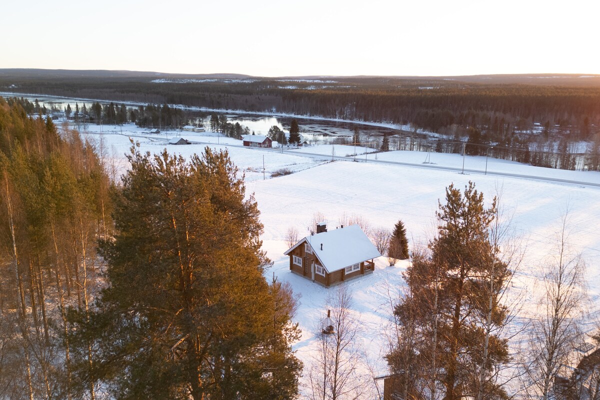 A cozy cottage is set amidst a snowy landscape, surrounded by tall pine trees. The area is tranquil, showcasing a vast expanse of white snow and a river in the distance, reflecting the warm light of a sunset.