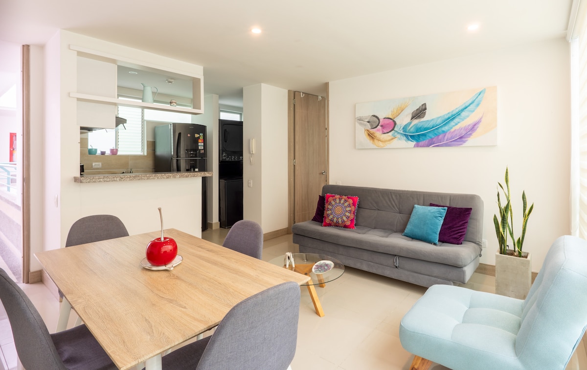 A modern living area is presented, featuring a gray sofa, a light blue armchair, and a wooden dining table. Bright artwork adorns the wall, while a decorative piece sits on the table. Large windows allow natural light to fill the space, enhancing its inviting ambiance.