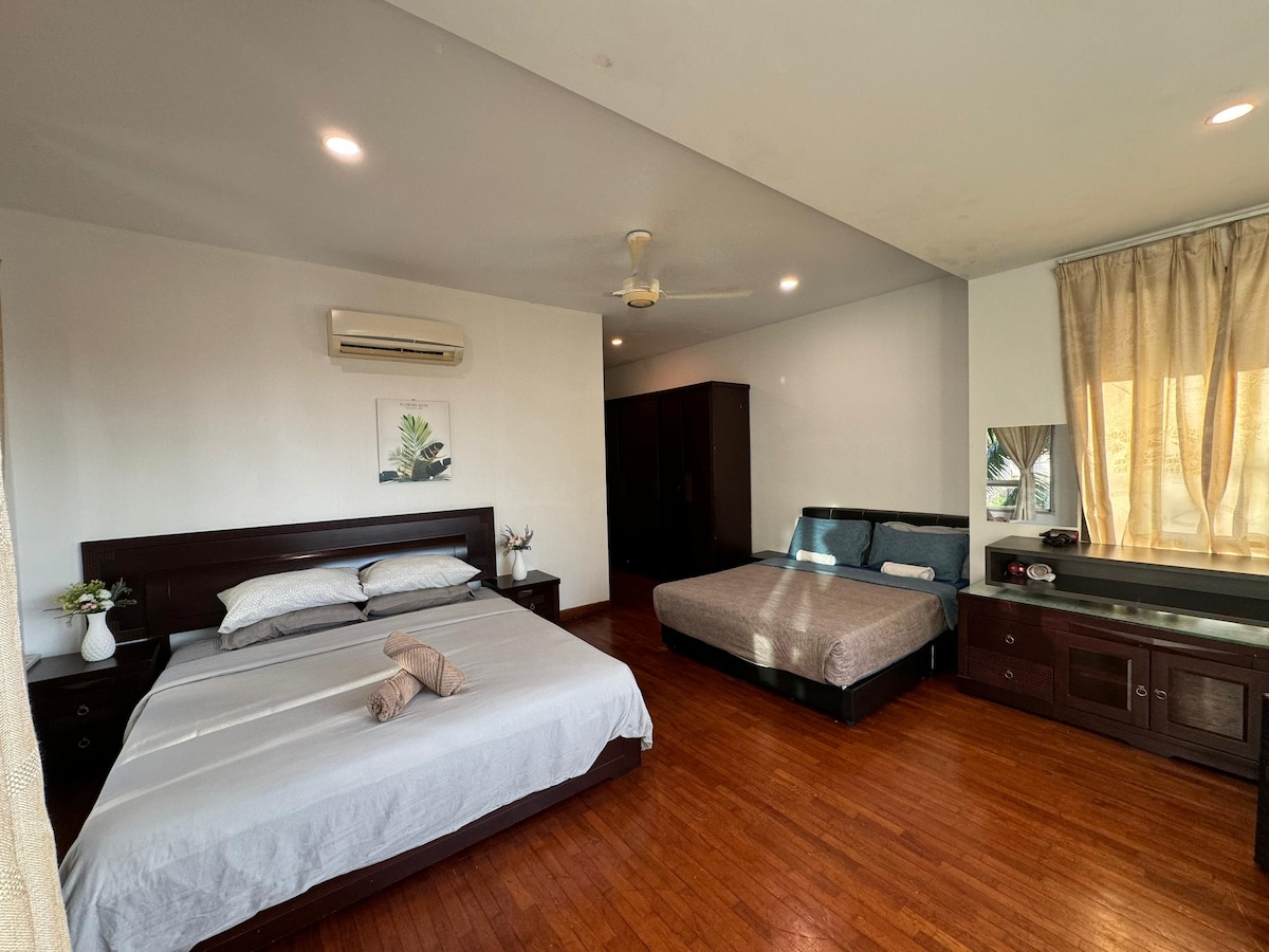 A spacious bedroom features two beds arranged thoughtfully, with one bed positioned against the wall. A wooden cabinet and a mirror are included in the setup. Soft natural light filters through the window, complemented by light-colored curtains and a plant decor on the side table.