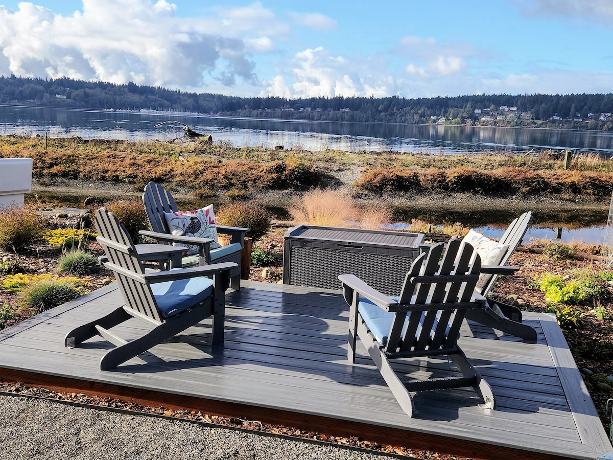 An outdoor seating area features four Adirondack chairs around a central fire pit, offering views of the tranquil water and surrounding landscape. Lush vegetation and natural elements frame the deck, creating a serene atmosphere for relaxation or gatherings.
