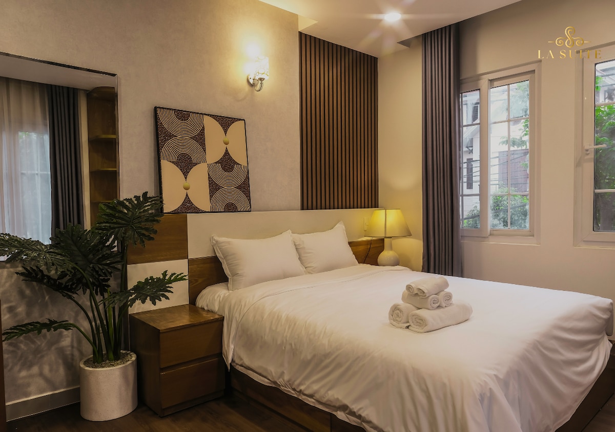A well-appointed bedroom features a spacious bed with neatly arranged towels. Soft lighting is provided by a bedside lamp, while a decorative wall art piece adds a subtle touch. Two windows allow natural light to fill the room, complemented by indoor plants near the mirror.