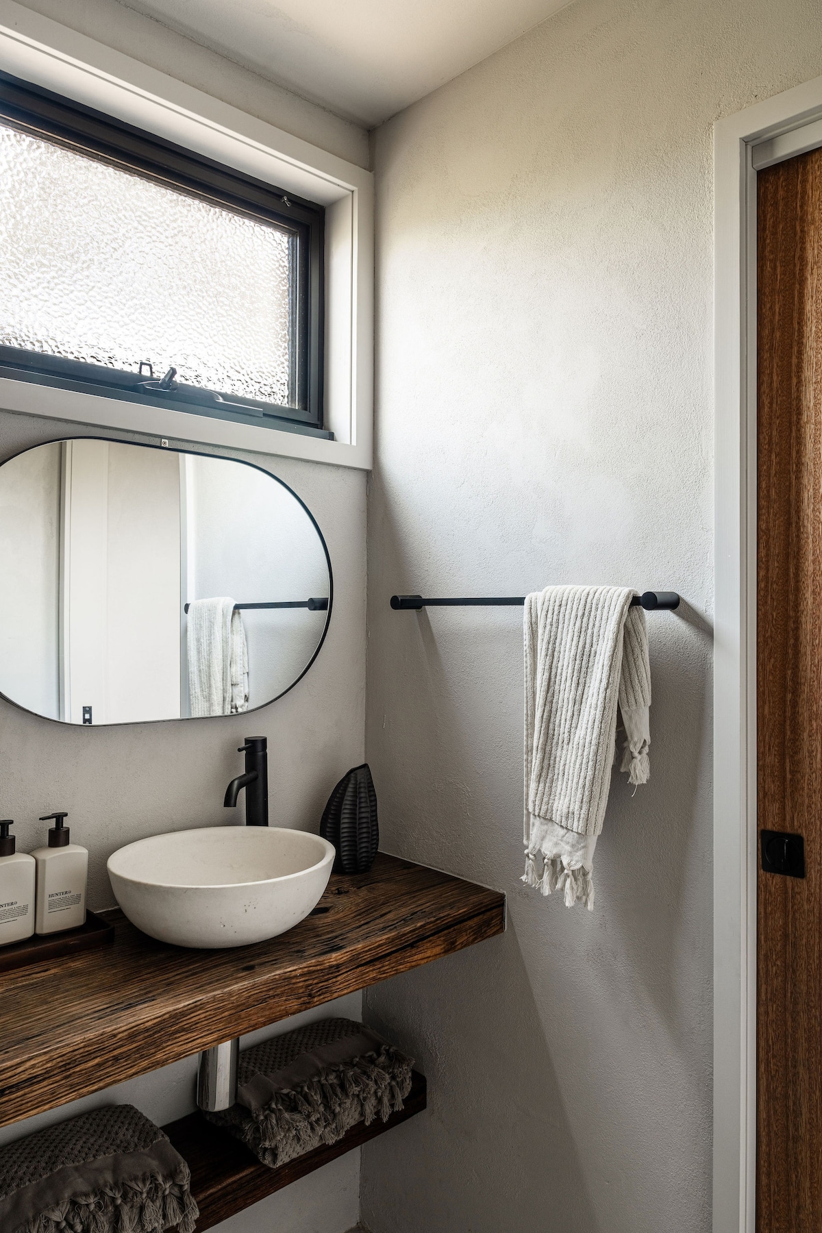 A modern bathroom features a circular mirror above a wooden countertop with a round basin. Natural light enters through a textured window. A black towel bar holds a folded towel, and neatly arranged toiletries are placed beside the basin on the countertop.