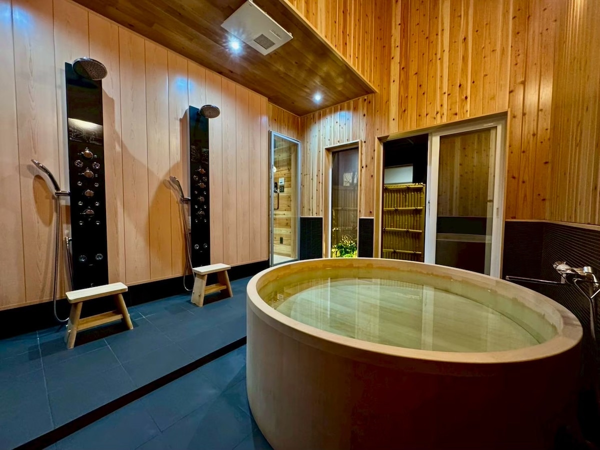 A serene bathing area features a circular hinoki wood bathtub surrounded by wooden paneling. Two shower stations are positioned nearby, complemented by stools for convenience. Soft overhead lighting enhances the tranquil atmosphere, while large windows provide a view into an adjacent space filled with greenery.