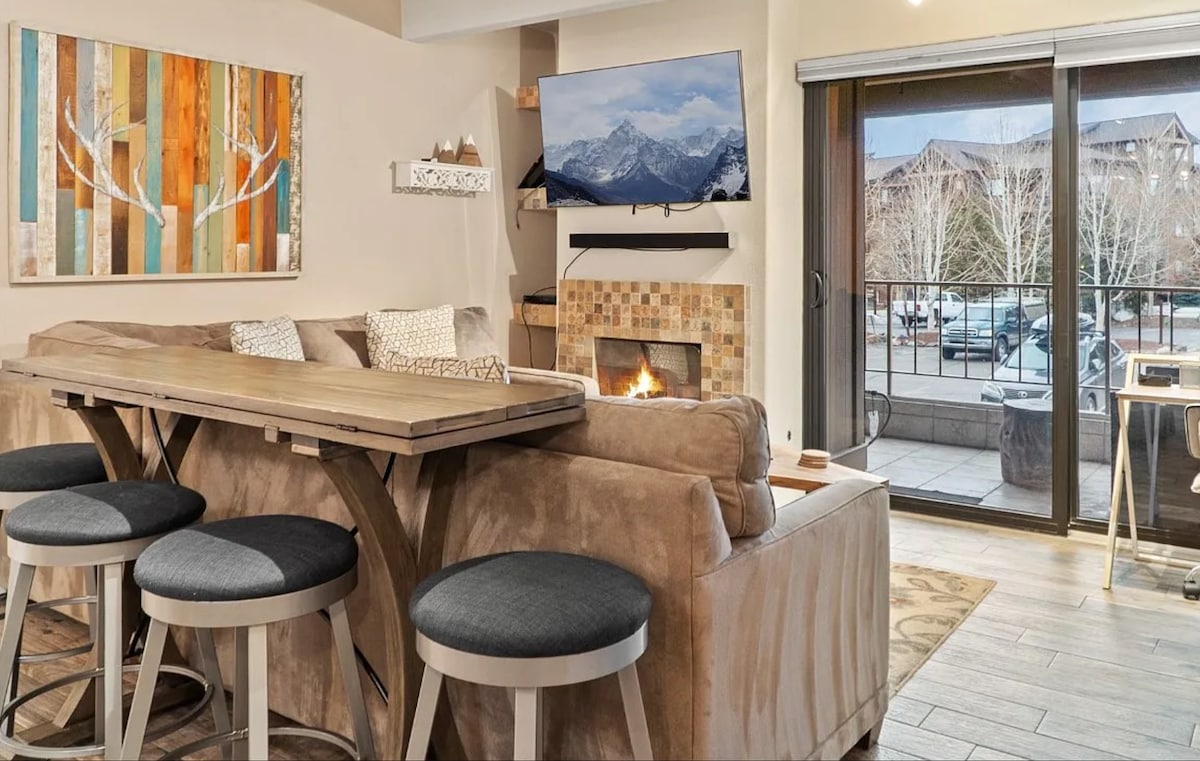 An open living area showcases a sectional sofa positioned near a fireplace. A large television is mounted on the wall, with a dining table featuring bar seating nearby. Sliding glass doors provide access to a patio with mountain views beyond.