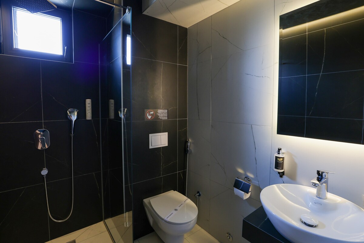 The bathroom features sleek black marble walls, complemented by modern fixtures. A spacious shower is enclosed with glass, and a circular sink is positioned beside a well-lit mirror. A minimalist toilet is situated against the wall, creating an airy ambiance.