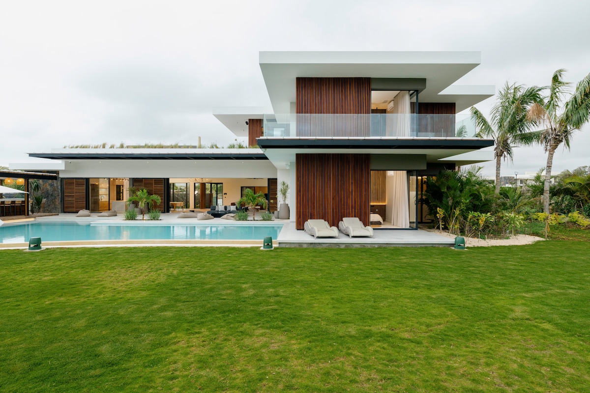 A contemporary villa is showcased with sleek lines, large windows, and wooden accents. The spacious lawn surrounds a rectangular pool, with several loungers positioned nearby. Tropical plants enhance the inviting outdoor space, creating a serene atmosphere against a cloudy sky.