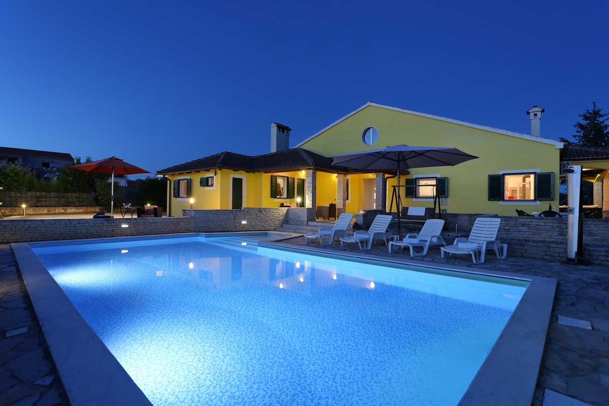 The villa is illuminated softly at dusk, showcasing a large outdoor swimming pool reflecting the evening sky. Surrounding the pool, multiple loungers are arranged for relaxation, and a sizable umbrella offers shade. The yellow exterior of the villa contrasts beautifully with the peaceful atmosphere.