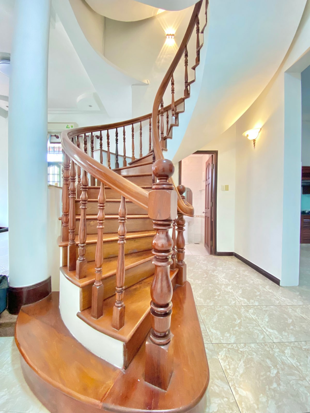 A gracefully curved wooden staircase is depicted, featuring polished railings and balusters. The steps, crafted from rich wood, lead upward, while soft lighting enhances the elegant ambiance of the space. White walls and marble flooring provide a clean backdrop, contributing to the overall refined aesthetic.