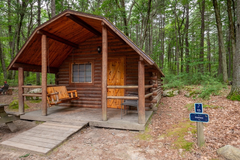 A rustic cabin features log construction with a pitched roof and a front porch. A wooden swing is positioned on the porch, accompanied by a small chair. Surrounded by tall trees, a sign indicating the cabin's name is visible in the foreground.