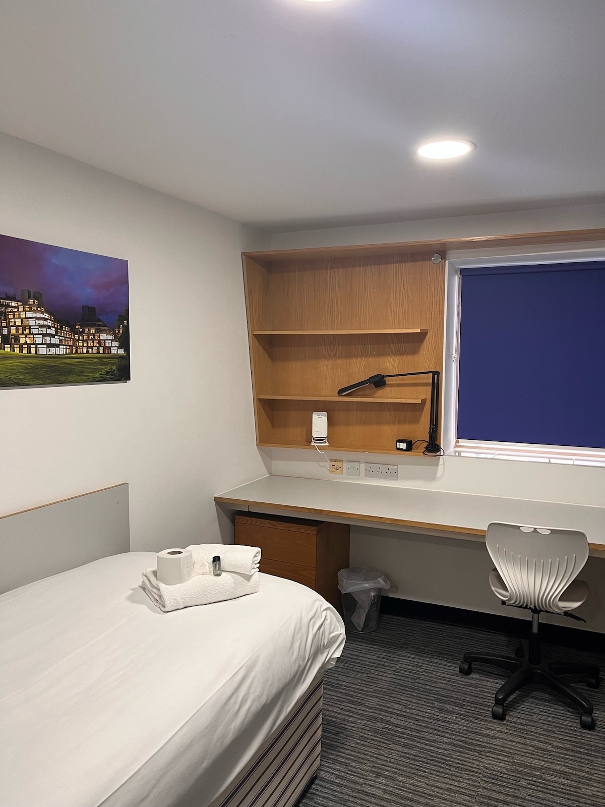 A single occupancy room features a comfortable bed with white linens and neatly folded towels. A compact workstation with a desk lamp is positioned next to a window with purple blinds, while a small shelving unit provides additional storage.