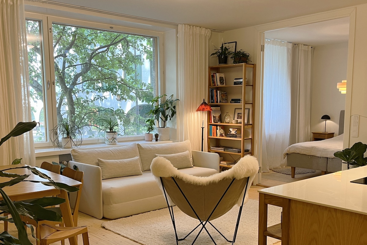An open-plan living area is shown, featuring a cozy sofa adorned with soft cushions, a relaxing chair with a light-colored fur throw, and natural light streaming through a large window overlooking greenery. A dining table and wooden bookshelf are visible, enhancing the inviting atmosphere.