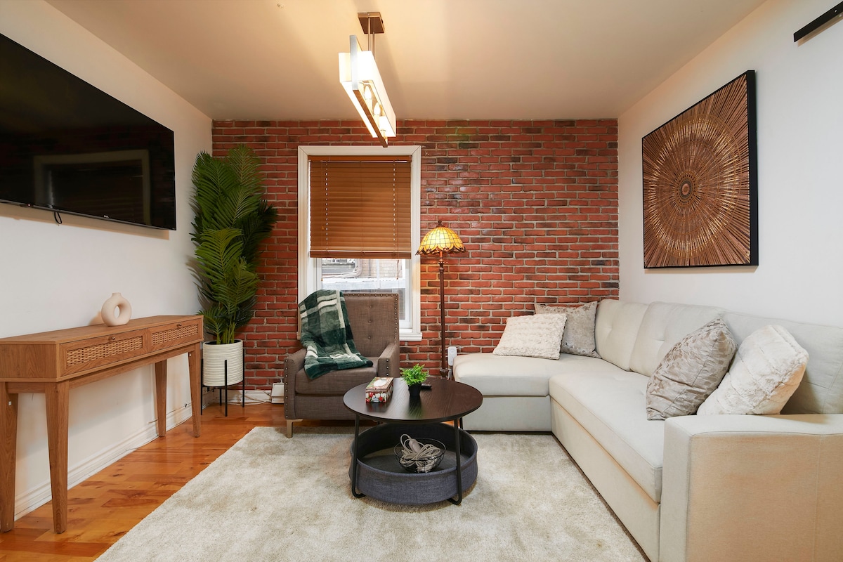 The living area features a contemporary sectional sofa alongside a cozy armchair, with warm lighting provided by a stylish lamp. A round coffee table sits at the center, surrounded by a plush area rug. Exposed brick walls add character, while large windows allow natural light to fill the space.