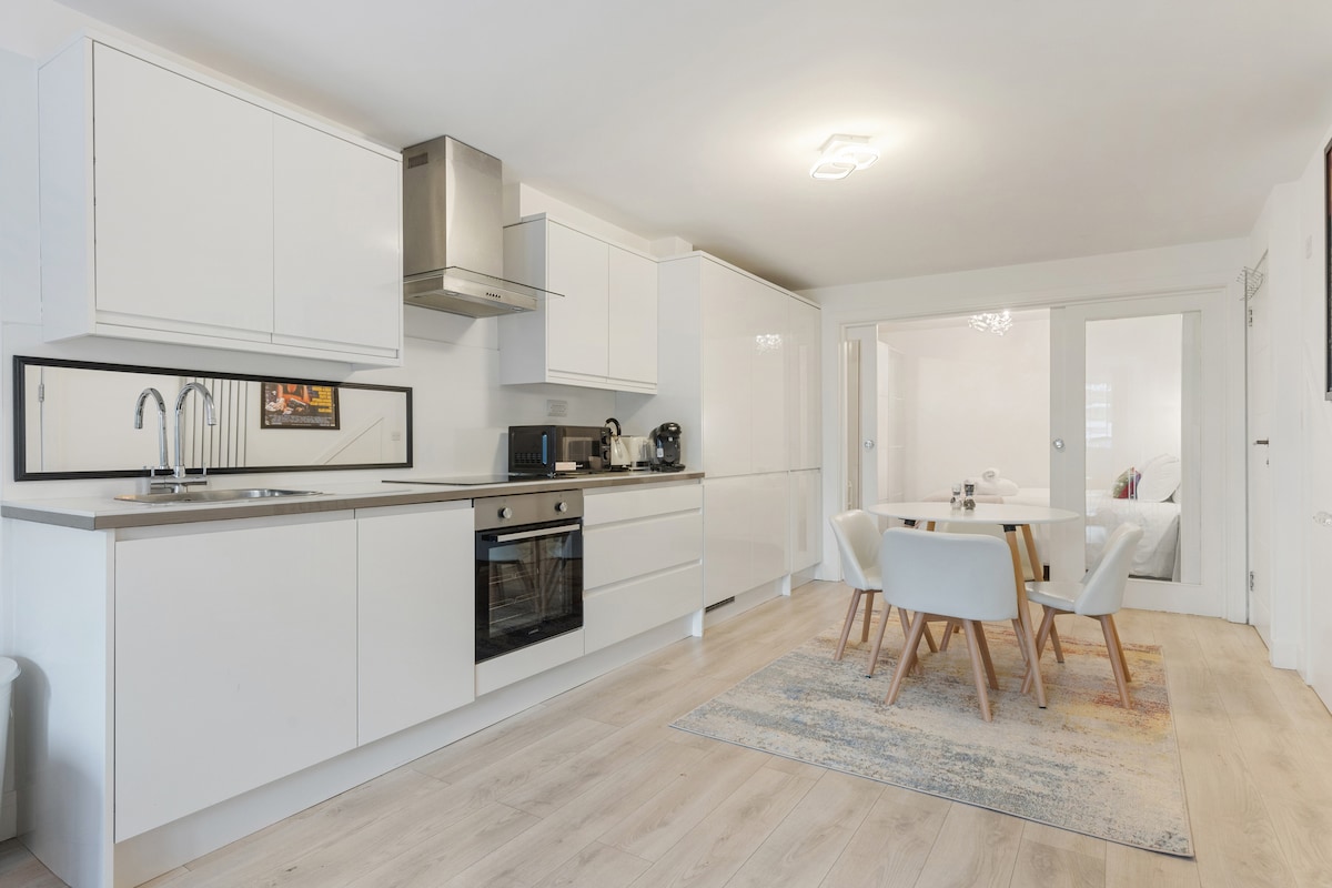 A modern kitchen and dining area is presented, featuring sleek white cabinetry and stainless steel appliances. A round dining table with four white chairs is positioned on a light-colored rug, creating an open and inviting space. Natural light fills the area, enhancing its brightness.