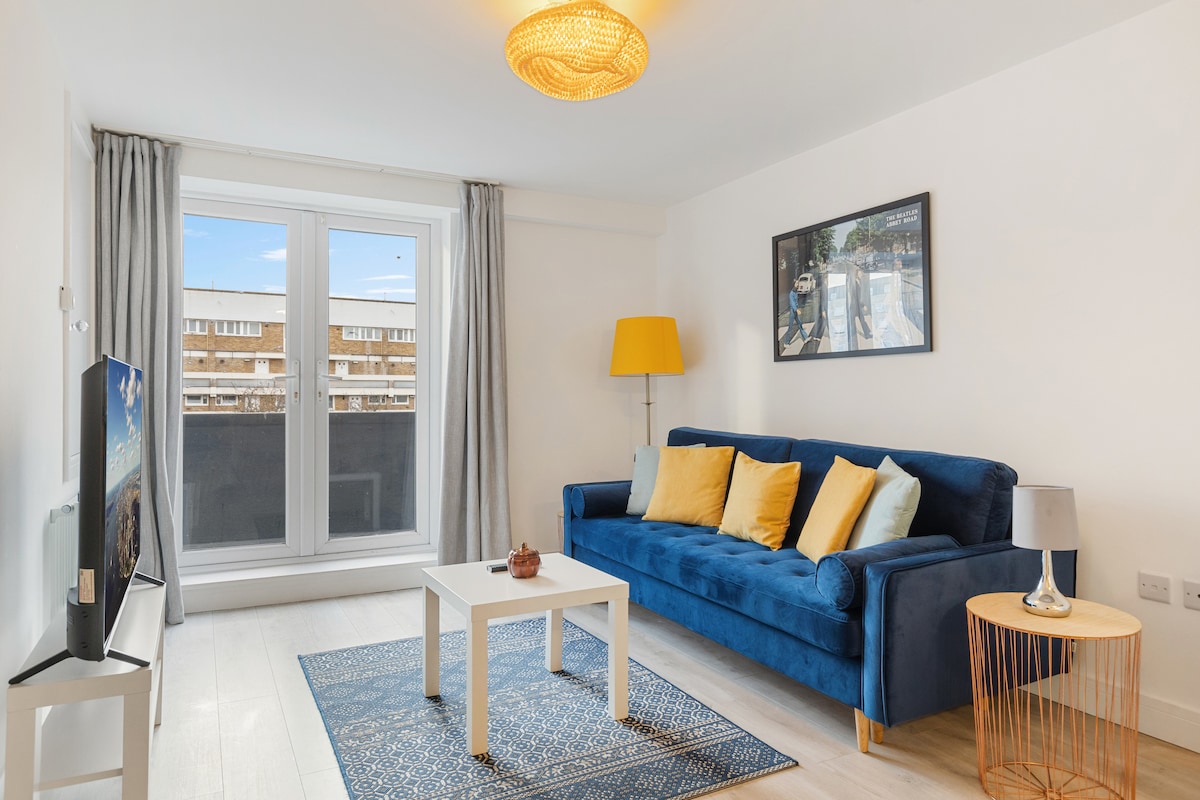 A cozy living area features a blue sofa adorned with yellow cushions, positioned beside a coffee table. A warm lamp stands near a window with sheer curtains, allowing natural light to brighten the space. A patterned rug adds texture to the light-colored floor.