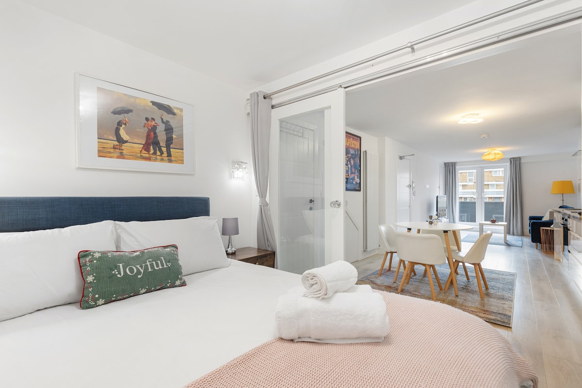 A spacious bedroom features a large bed adorned with a decorative throw pillow and neatly rolled towels. An artistic print hangs above the bed. The open layout reveals a dining area with a modern table and chairs, visible through a glass partition.