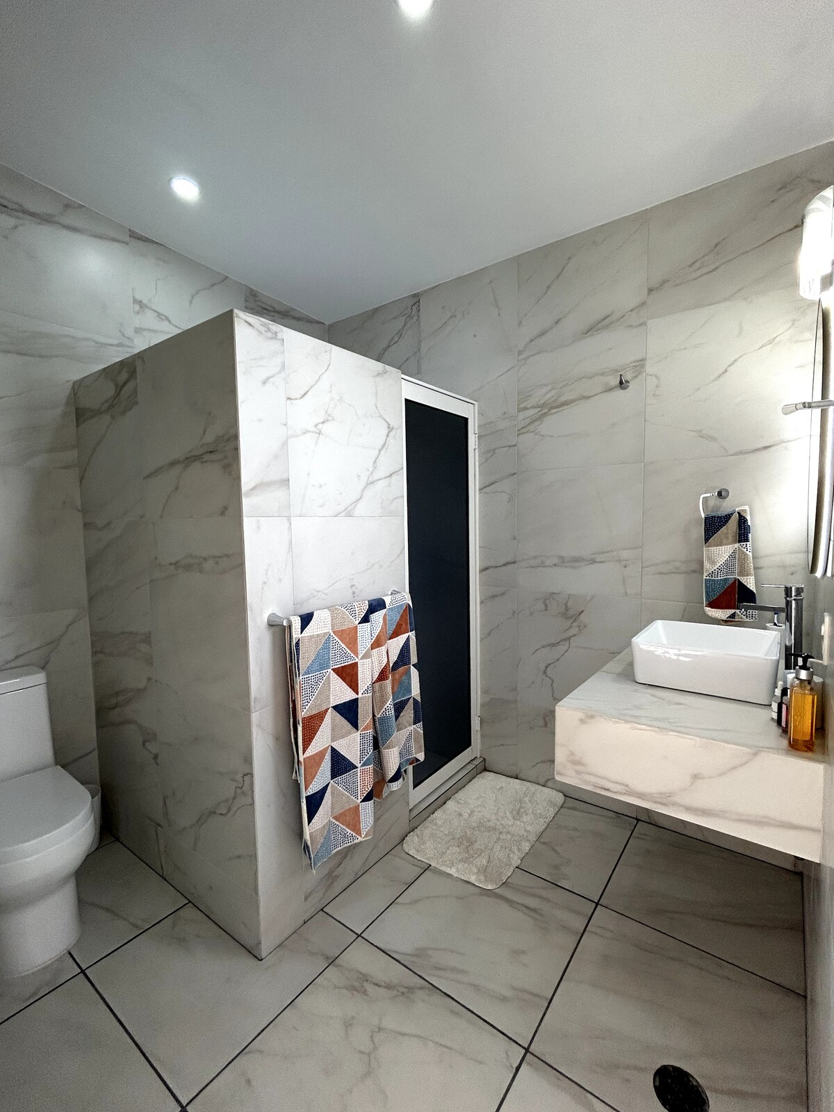 The modern bathroom features elegant marble walls, a spacious shower area separated by a glass door, and a sleek vanity with a white basin. Colorful towels hang decoratively, and the floor is tiled in a light hue, creating a clean and contemporary look.
