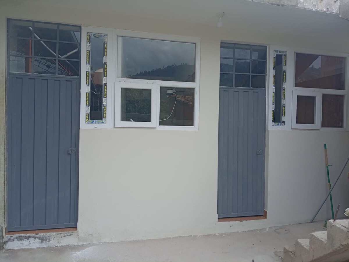 Two grey doors are framed by large windows in a light-colored exterior. The doors lead into separate rooms, while the windows allow natural light to enter. A muted background of green hills is visible beyond the windows.
