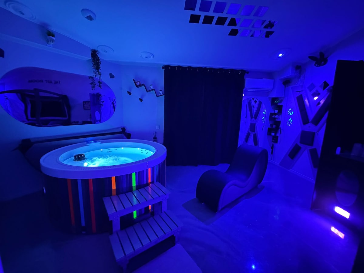 A stylish room is enhanced by colorful lighting, featuring a round luxury spa and a modern lounge chair. A large mirror and curtains add depth, while the unique decor on the walls invites exploration. Soft lighting casts a tranquil ambiance throughout the space.
