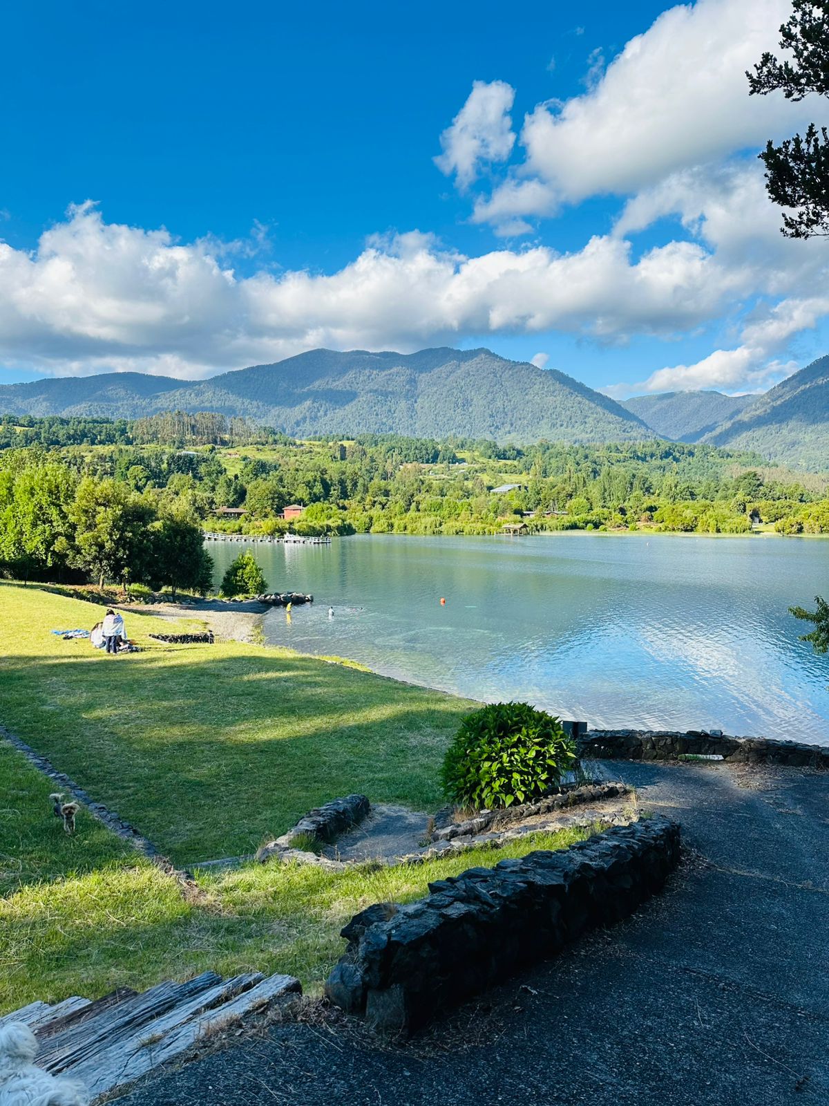 A scenic view of Lago Ranco is presented, framed by rolling green hills and mountains under a blue sky scattered with fluffy clouds. A pathway leads down to the water's edge, where individuals enjoy the tranquil surroundings, surrounded by vibrant foliage.