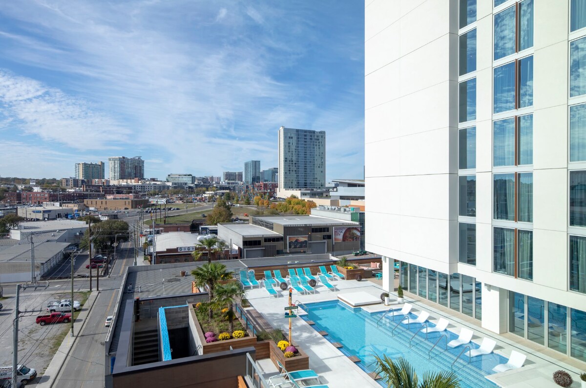 An expansive view showcases the rooftop pool area, flanked by colorful lounge chairs and tropical landscaping. The city skyline is visible in the background under a clear blue sky, creating a bright and modern atmosphere.