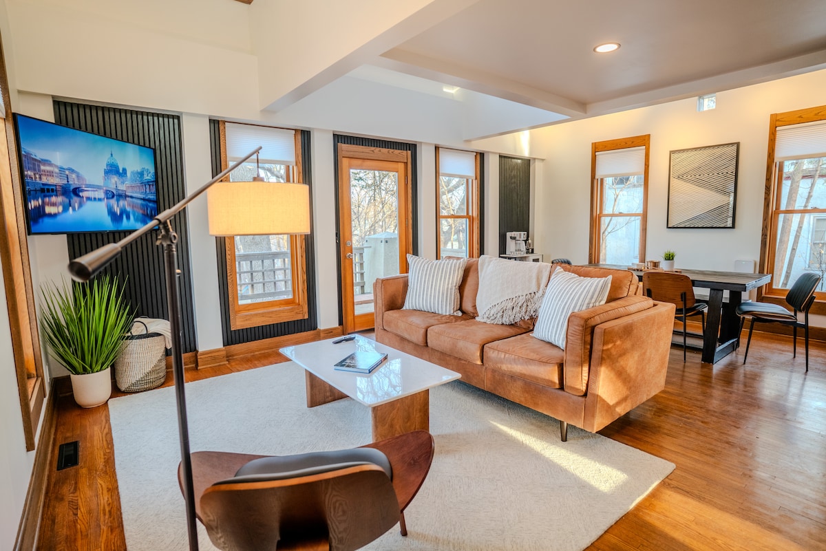 A modern living area is highlighted by a large sectional sofa with soft cushions, accompanied by a sleek coffee table. Natural light streams through multiple windows, illuminating the space. A contemporary lamp stands nearby, and a dining area with simple chairs is visible in the background.