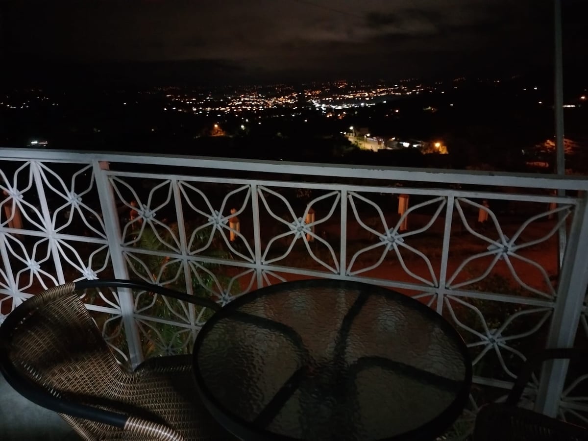 A balcony scene reveals a glass table accompanied by two woven chairs, surrounded by intricate railings. The night view showcases a distant city illuminated by lights, creating a serene backdrop against the dark sky.