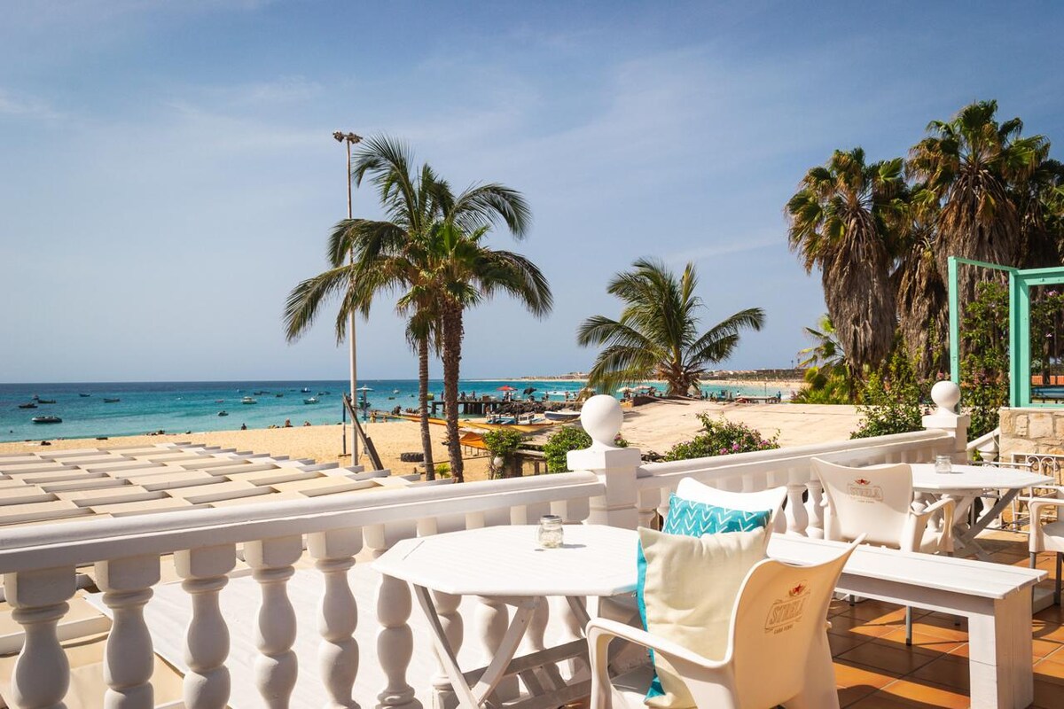 A shared terrace features white furniture positioned for ocean views, with palm trees and sandy beach visible in the foreground. The azure sea sparkles under a clear blue sky, while groups of small boats are seen on the water, enhancing the coastal ambiance.