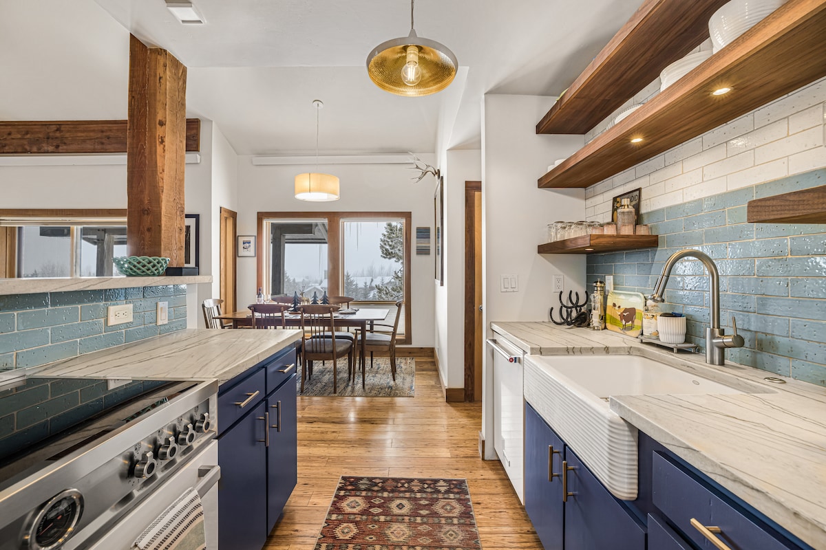 The kitchen features blue cabinetry with marble countertops and a farmhouse sink. Overhead, a pendant light provides illumination. A dining area is visible in the background, framed by large windows offering views of the landscape beyond. Hardwood floors add warmth to the space.