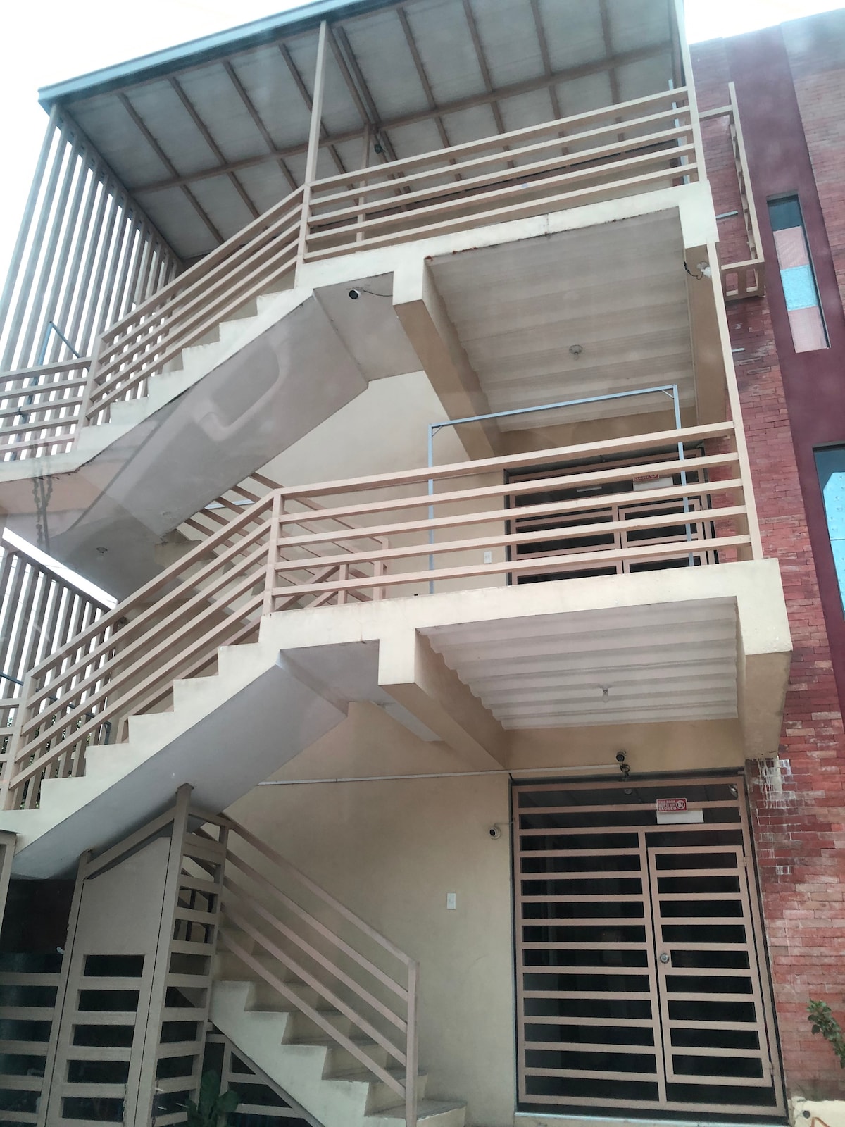 A three-story building is depicted, showcasing a modern exterior design with multiple staircases. Each flight of stairs features open railings, allowing for easy access to the upper levels. A doorway is visible on the lower level, framed with simple architectural lines.