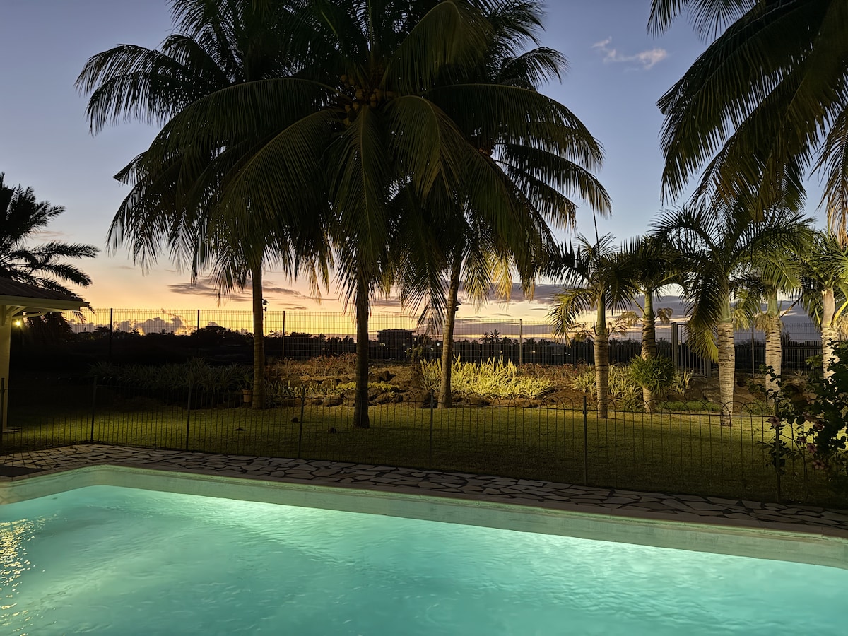 A serene view of the sparkling pool illuminated at dusk, surrounded by lush palm trees. The vibrant colors of the sunset reflect on the water, creating a tranquil atmosphere in the spacious garden. A gentle breeze can be imagined rustling through the foliage.