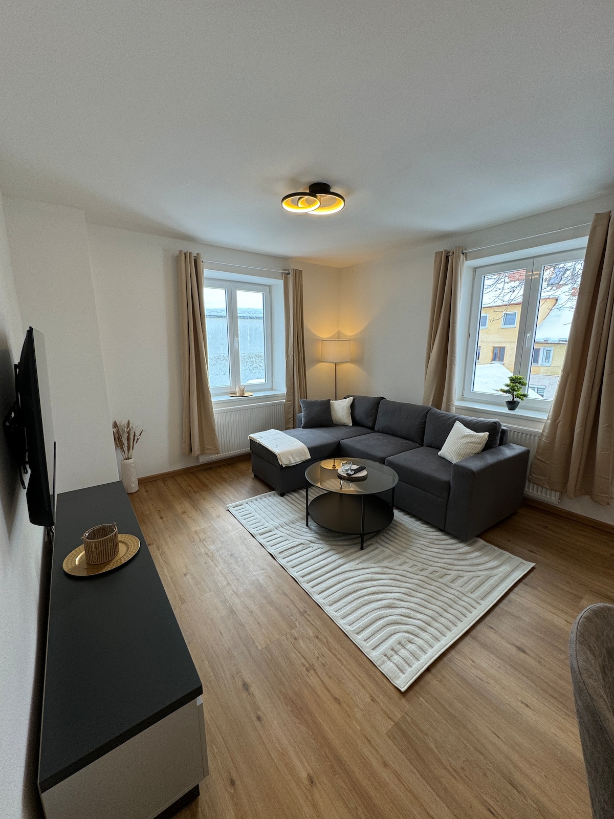 A modern living area is presented, featuring a comfortable gray corner sofa paired with a round coffee table. Natural light enters through large windows adorned with light curtains. A light rug enhances the warmth of the wooden floor, while decorative elements add subtle detail.