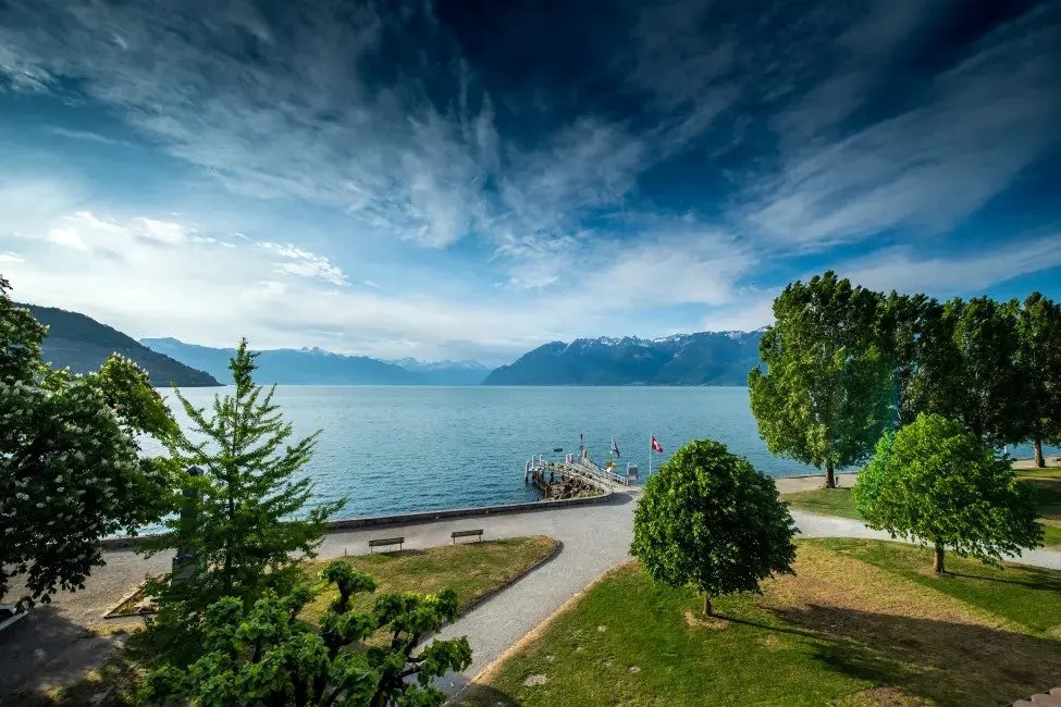 A serene view of the lake, featuring calm waters and distant mountains under a partly cloudy sky. Lush green trees line the shores, with a pathway leading to a small pier. Benches provide a space for relaxation near the water's edge.