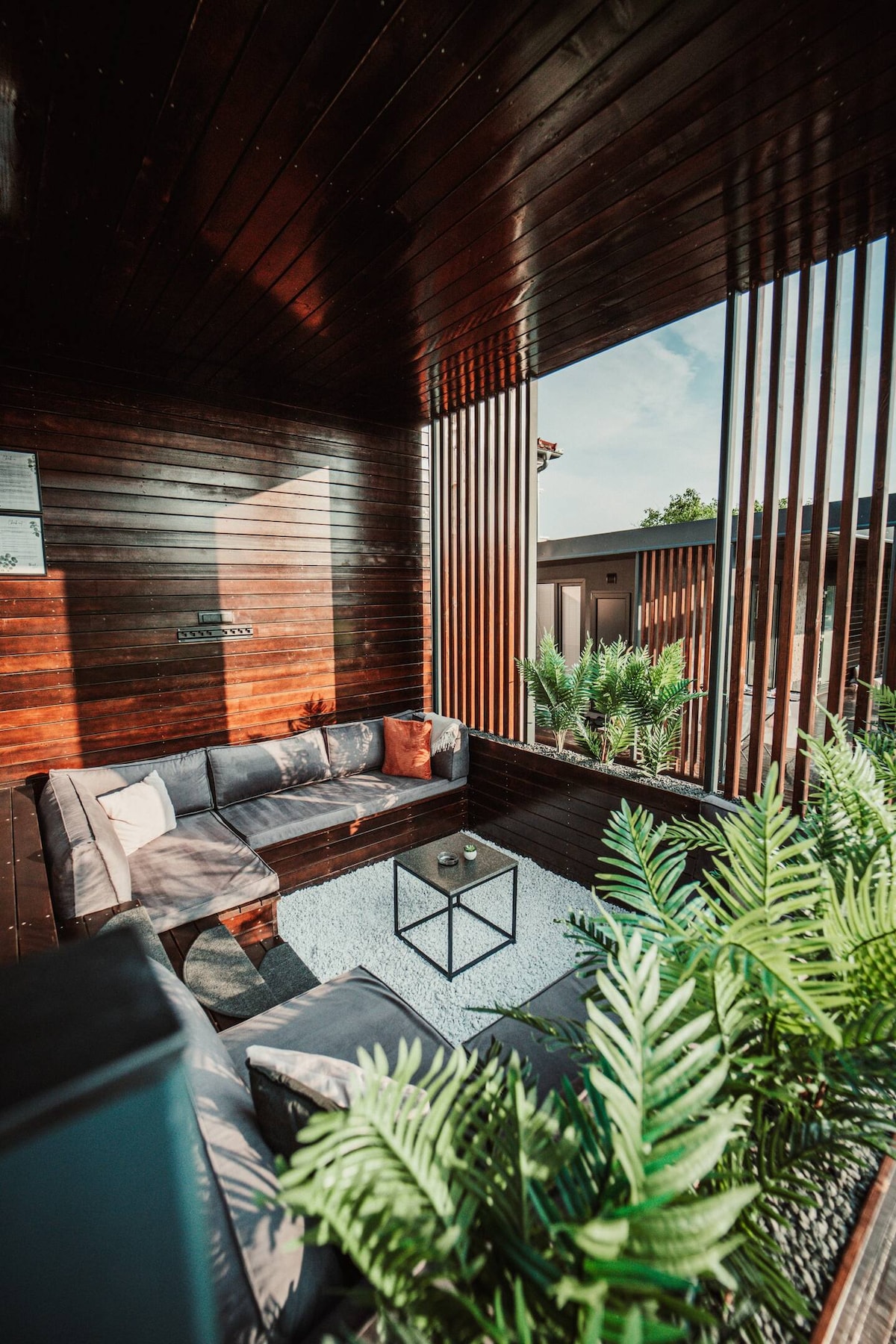 A shaded outdoor seating area is surrounded by wooden paneling and greenery. Comfortable couches are arranged around a glass-top table, resting on a white gravel floor. Vertical slats provide a sense of privacy while allowing natural light to filter through.
