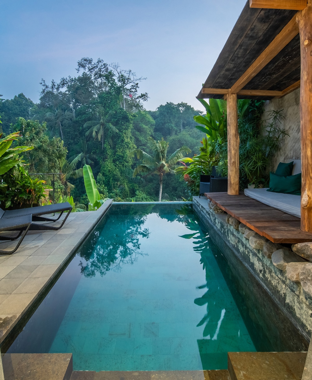 A private pool reflects the surrounding lush jungle, framed by natural stone walls and wooden beams. Comfortable lounge chairs are positioned nearby, while tropical plants enhance the serene atmosphere. The scene conveys tranquility, inviting relaxation amid the vibrant greenery.