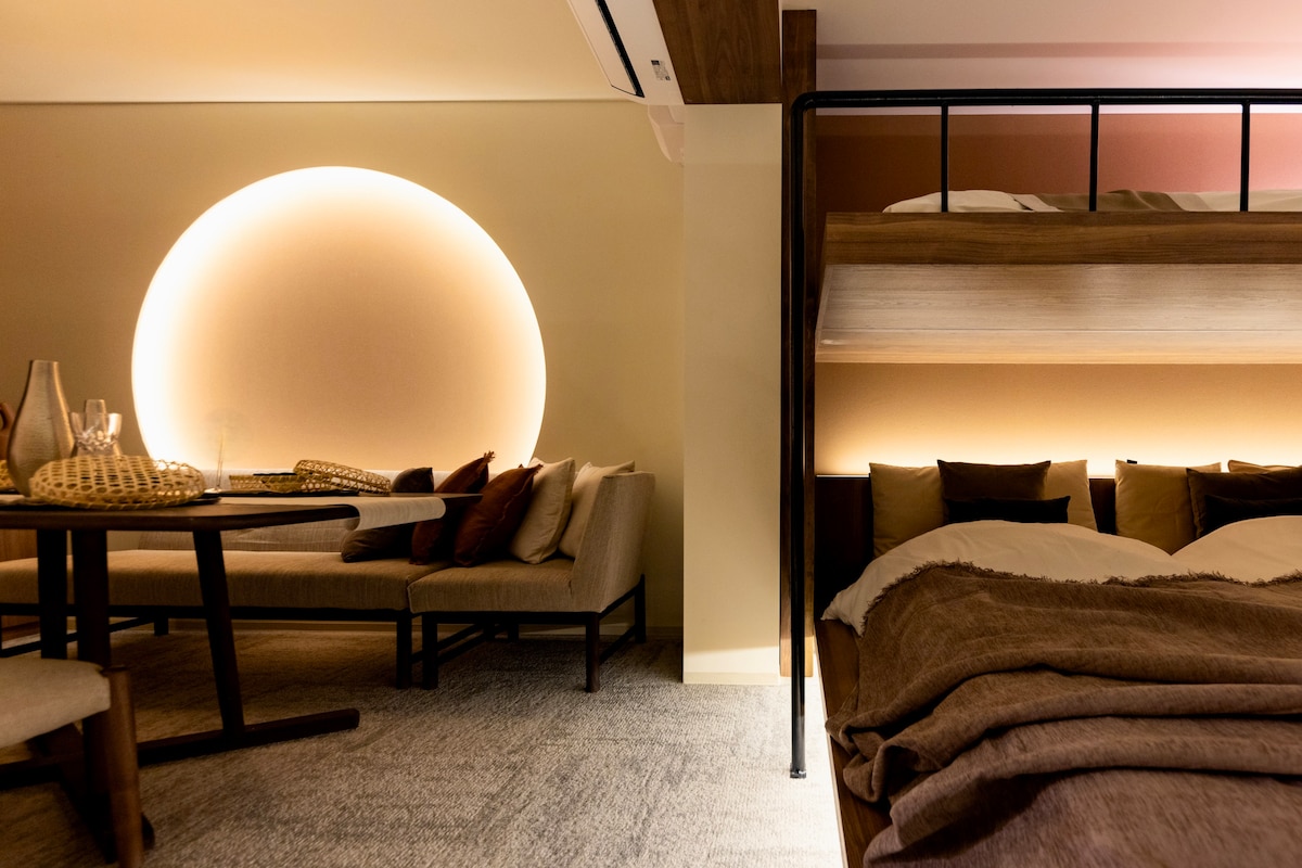 A cozy interior features a large circular light installation that creates a warm ambiance. The space includes a bunk bed with plush bedding and a seating area with a stylish table, accommodating comfortable dining and relaxation.