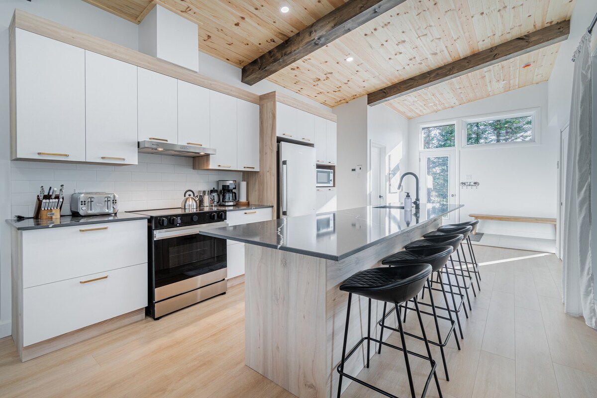 A modern kitchen features a spacious central island with seating for four. Stainless steel appliances complement sleek cabinetry, while wooden beams enhance the ceiling's height. Natural light fills the room through large windows, creating a bright and inviting cooking space.