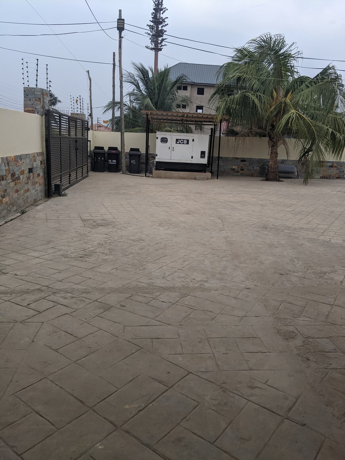 The outdoor area features a spacious, paved surface with a few palm trees in view. A backup generator is located under a covered structure, while a gated entrance is visible in the background. Several bins are arranged neatly along the side.