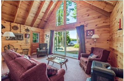 A cozy living space features wooden walls and a high, sloped ceiling. Two comfortable armchairs and a sofa are arranged for relaxation. Large glass doors allow natural light to fill the room and provide a view of the lake outside, enhancing the home's connection to nature.
