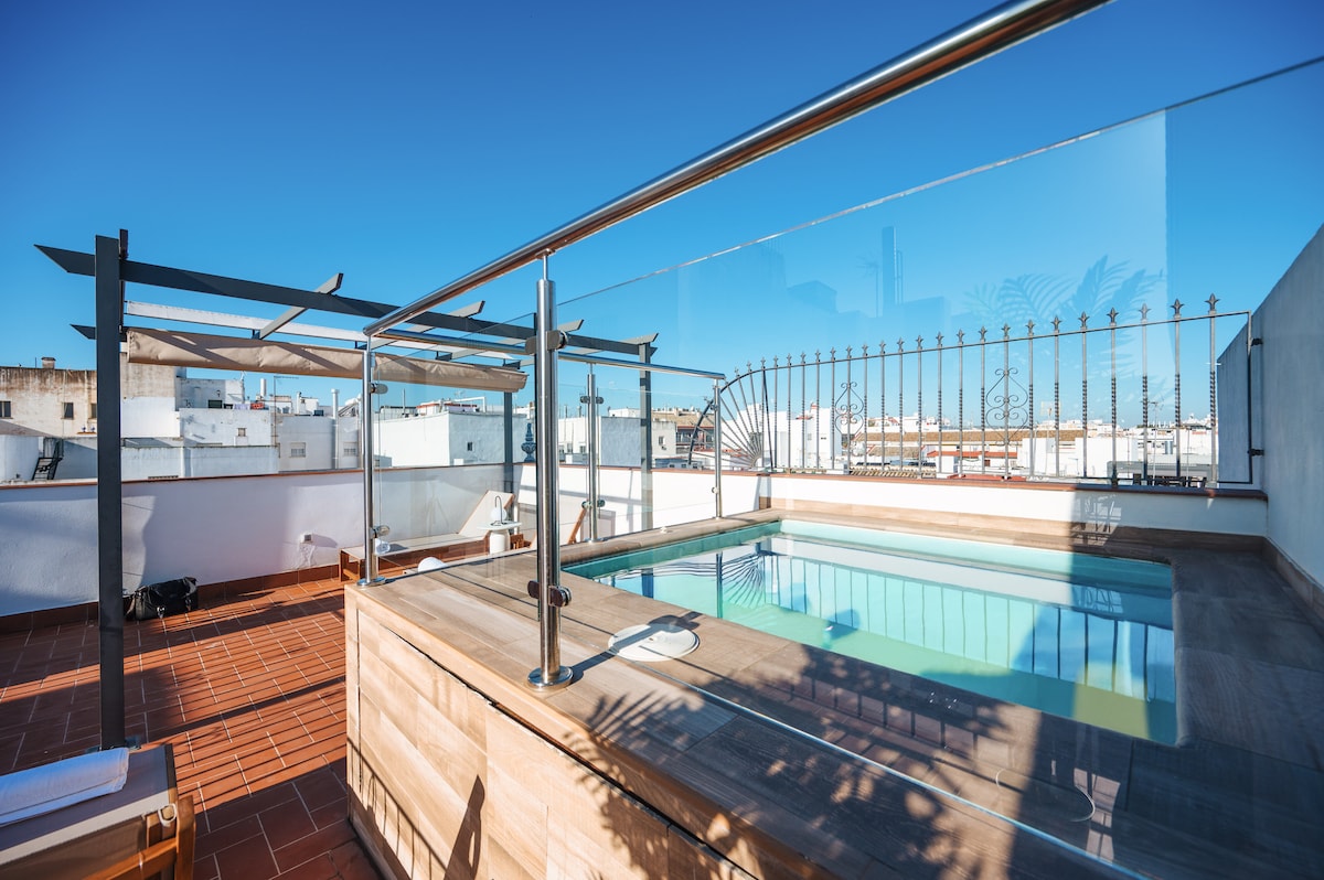 A bright rooftop area is highlighted by a clear glass barrier surrounding a small pool. Comfortable lounge chairs are positioned nearby on a tile deck, providing an inviting space for relaxation under a clear blue sky.