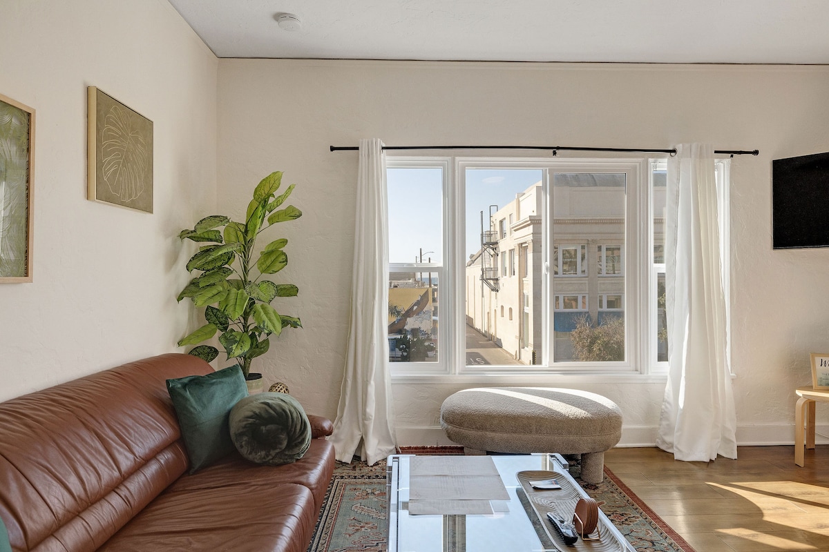 A cozy living area is presented, featuring a leather sofa complemented by decorative pillows. A large window offers a view of the surroundings, adorned with sheer white curtains. A round coffee table sits in front, with a small accent chair nearby, creating a comfortable and inviting space.