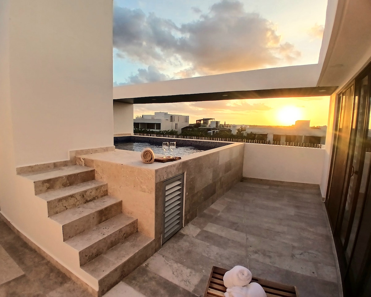 A serene rooftop terrace features a private pool, with steps leading up from the surface. Lounge chairs are positioned nearby, offering a view of the sunset over Tulum. The space is surrounded by a clean architectural design, enhancing the tranquil atmosphere.