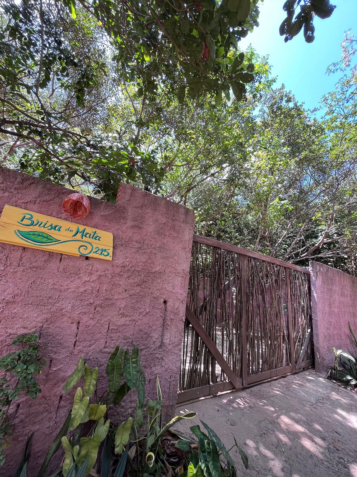 An entrance gate made of wood is framed by natural bamboo fencing, set against a textured pink wall. Lush green foliage surrounds the area, with a colorful wooden sign displaying 'Brisa da Mata 225' attached to the wall, indicating the welcoming entryway.