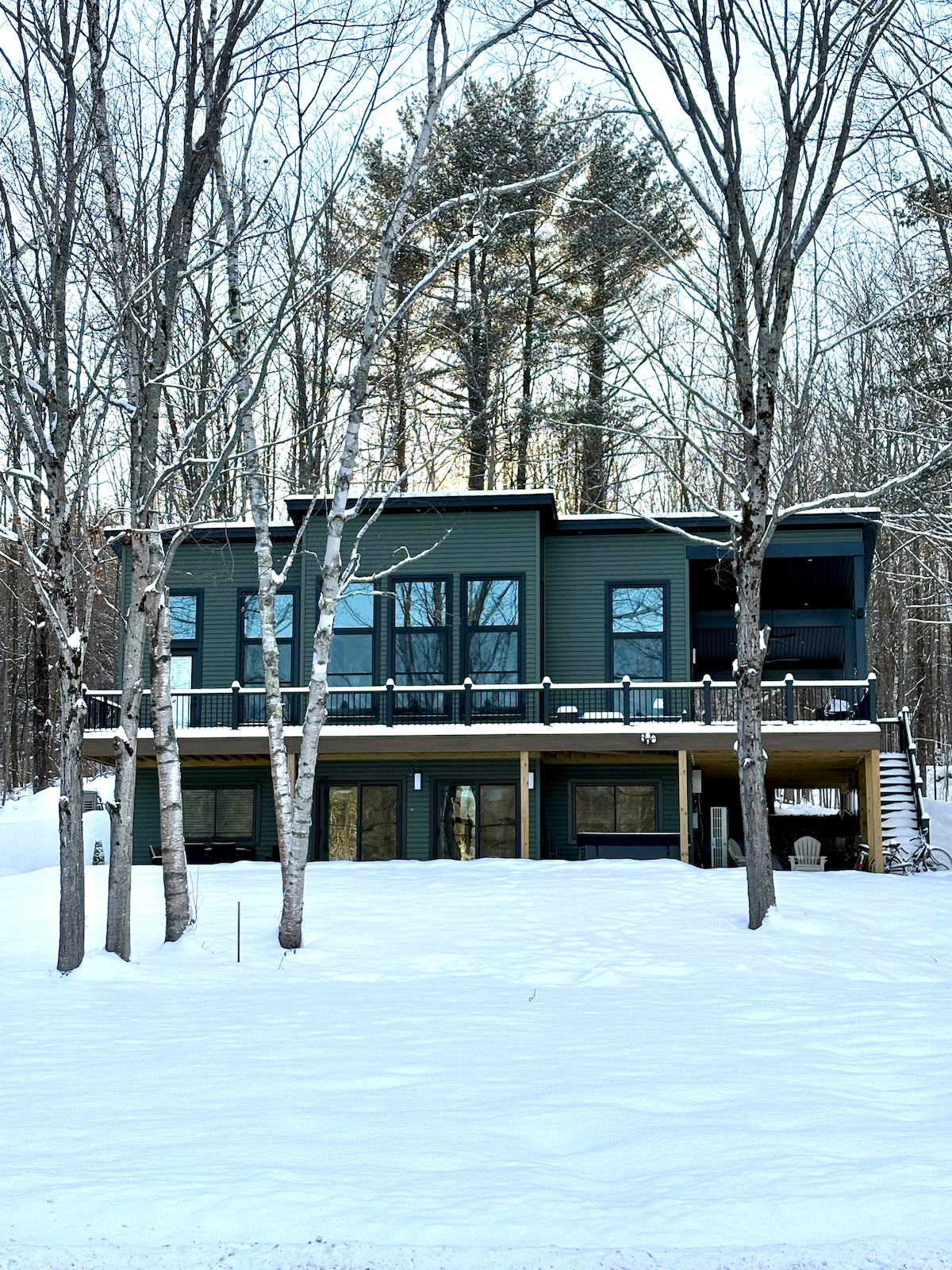 The exterior of the contemporary retreat is surrounded by snow-covered ground, showcasing large windows and a wraparound deck. Two levels are visible, with the upper part featuring a covered porch, while the lower level displays ample glass doors, inviting natural light into the home.
