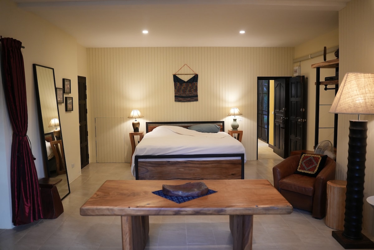 A serene bedroom space is presented, featuring a spacious bed centered in the room. A wooden table is positioned nearby, complemented by a leather chair. Soft lighting from side lamps creates a calming ambiance, while doors lead to the attached bathroom and outdoor areas.