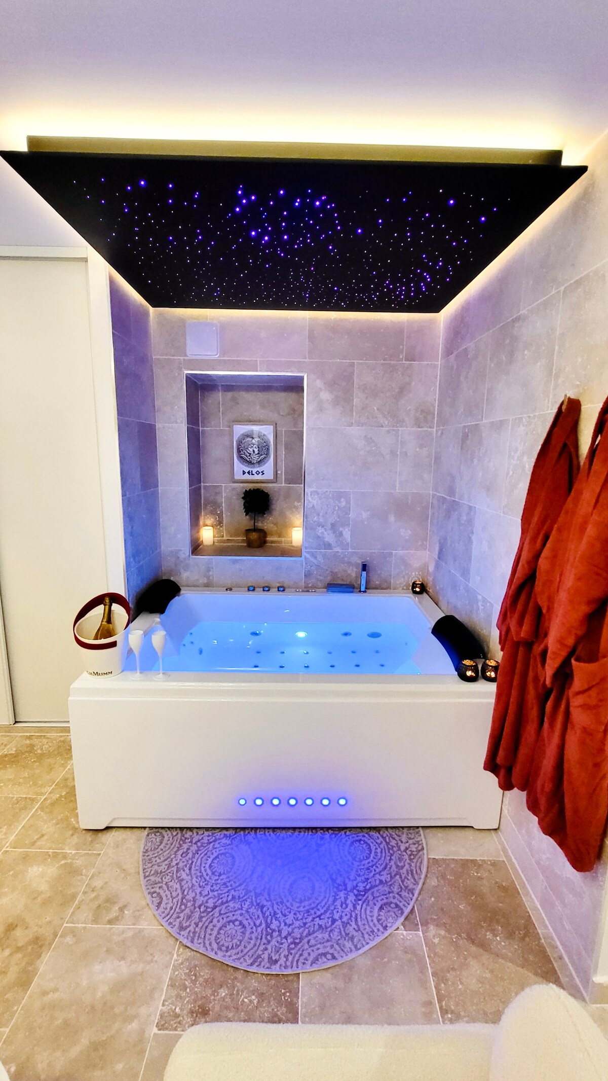 A spacious bathroom is highlighted by a large whirlpool bathtub designed for two. Soft blue lighting enhances the atmosphere, while the ceiling features a starry night design. Plush bathrobes are hung nearby, complementing the elegant tile work and modern fixtures.