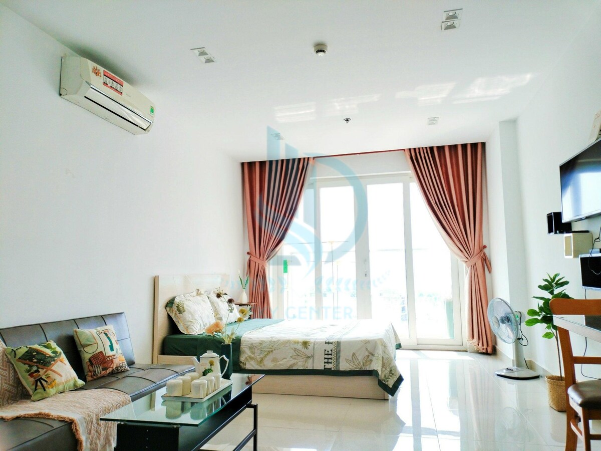 A contemporary living space features a spacious bed and a comfortable sofa. Light-colored curtains frame the large glass doors, allowing natural light to fill the room. A small fan and a wooden shelf are also present, contributing to an organized layout.