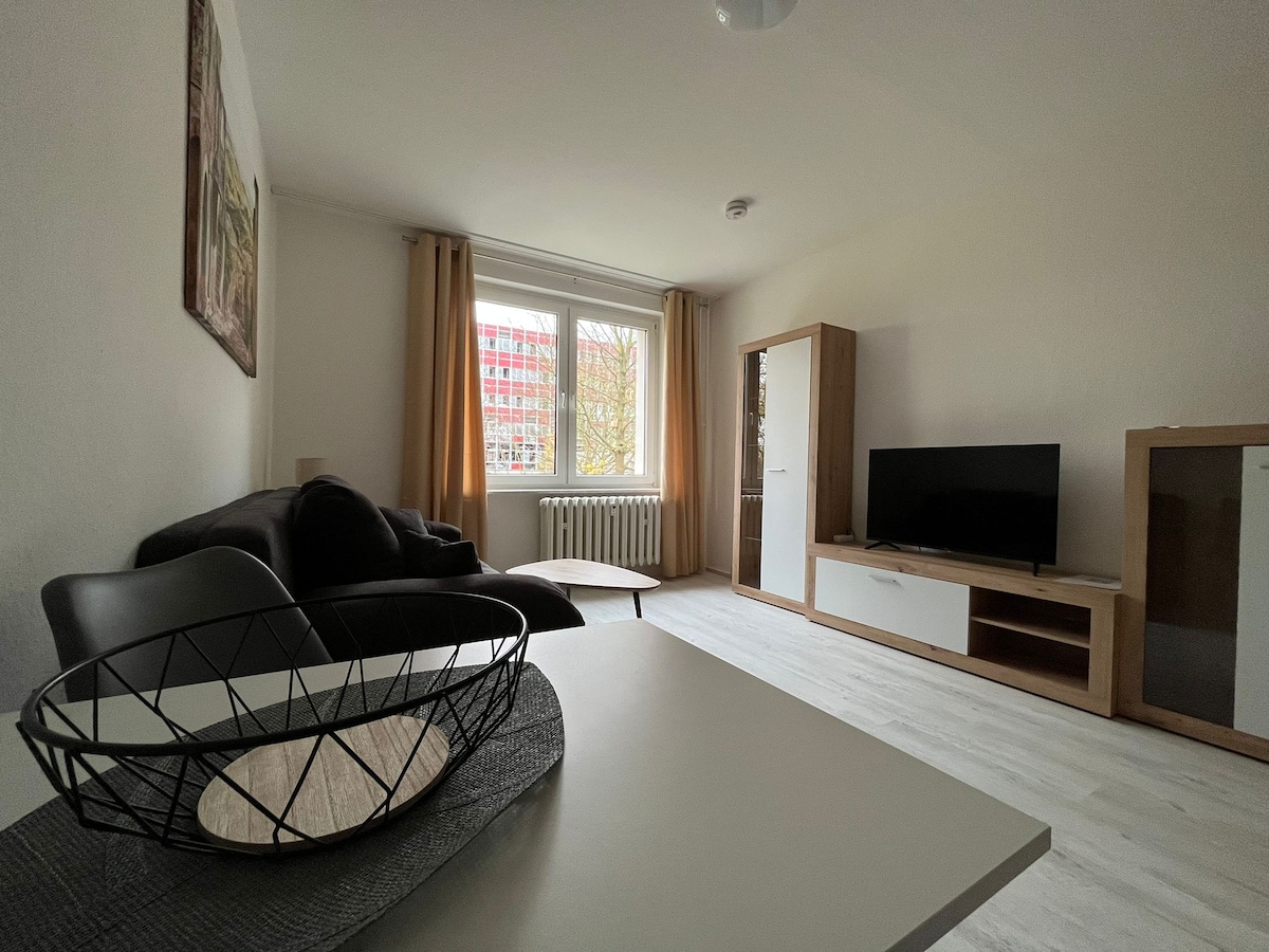 A modern living area features a comfortable black sofa and a light-colored coffee table. Natural light is captured through a large window covered by light curtains. A television is mounted on a unit beside a wooden shelving cabinet, providing ample storage space.