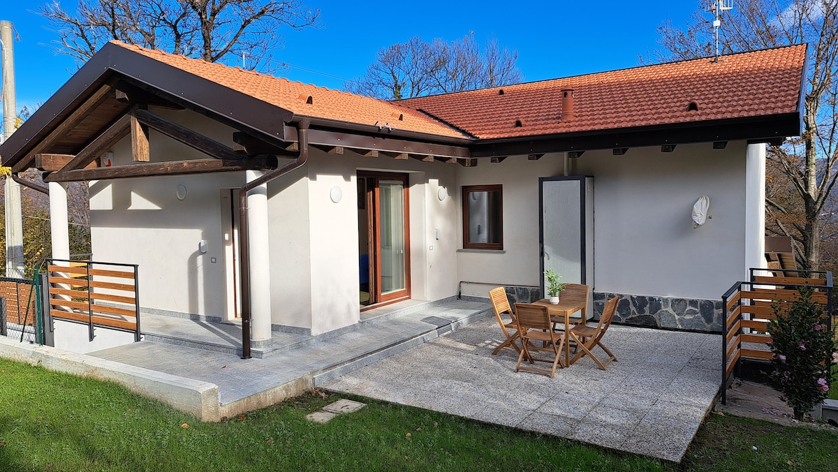 A modern exterior of the independent unit is displayed, featuring a spacious terrace furnished with a small round table and four chairs. The building is complemented by a sloped roof and is surrounded by green grass and trees, creating a serene outdoor space.