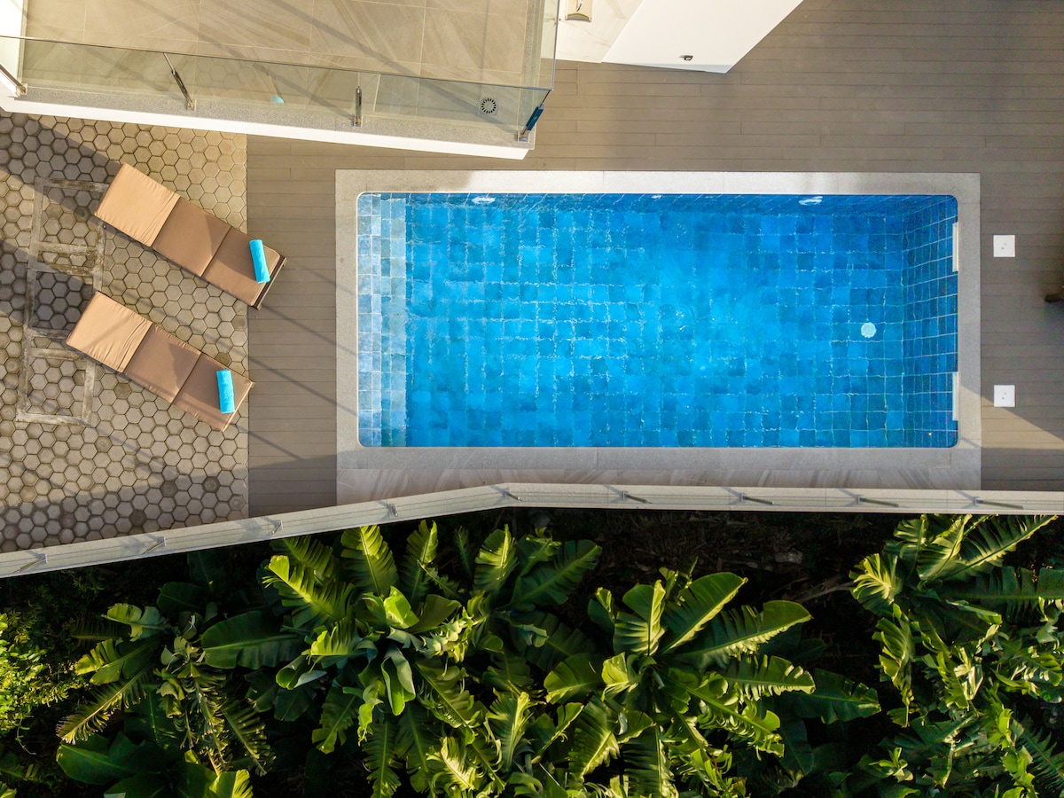The outdoor space features a modern pool surrounded by lush greenery, with two sun loungers positioned nearby. The pool's clear blue water invites relaxation, while the surrounding plants enhance the natural setting.